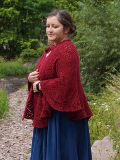 Side view of large red Ruby phoenix capelet Tunisian crochet sample worn over the shoulders, with the wavy bottom draped over partly folded arms.