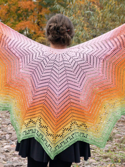 Crochet shawl made from a vest with the armholes pinned close, with a gradient color transition from soft pink to warm orange and vibrant green. The intricate lace detailing on the outer edge features a seamless geometric pattern.